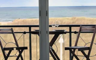 Balkon mit Tisch, zwei Stühlen, Blick auf die Ostsee, Dünen und Strand durch die geöffnete Balkontür.