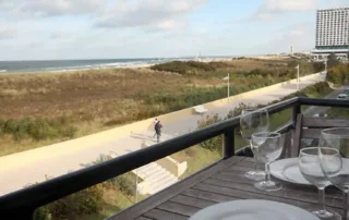 Balkon mit eingedecktem Tisch, Tellern, Gläsern und Aussicht auf Promenade, Dünen und Ostsee.