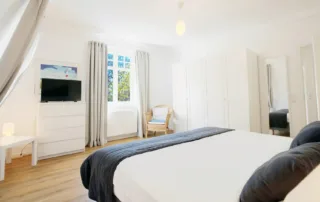 Schlafzimmer mit Bett, Fernseher, Fenster, Kommode, Spiegel und Kleiderschrank in heller Einrichtung